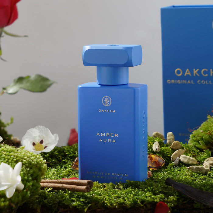 Front view of Amber Aura perfume bottle in matte cobalt blue with rectangular cap and gold logo on moss and floral background