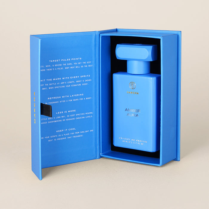 Front view of Amber Aura 50ml perfume bottle in blue presentation box showing matte blue bottle and fold-open packaging on beige background