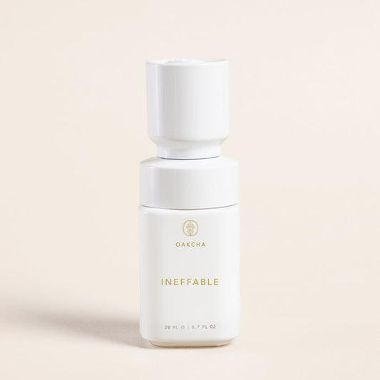 Front view of Ineffable Perfume Oil bottle showing a minimalist white bottle with gold logo on a light beige background