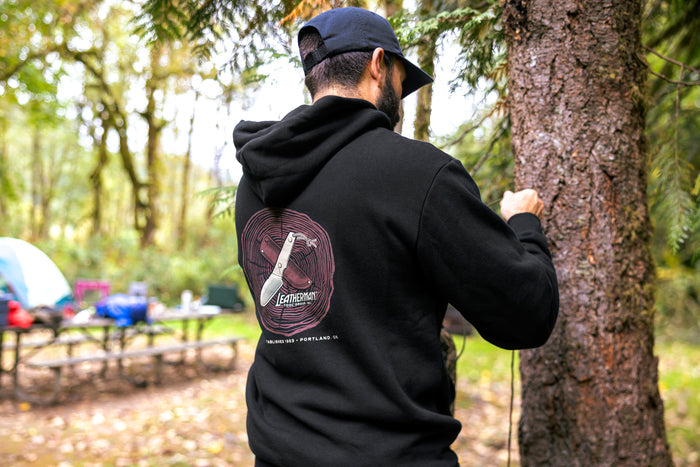 A person wearing a black Leatherman Blade & Grain hoodie standing next to a tree in a forest campsite. The hoodie’s back graphic shows a pocketknife illustration with “Leatherman Tool Group” text.