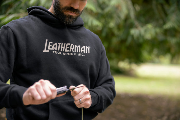 A person wearing a black Leatherman Blade & Grain hoodie using a Leatherman multitool to cut a piece of rope outdoors, showing the front logo of the hoodie that reads “Leatherman Tool Group, Inc.”