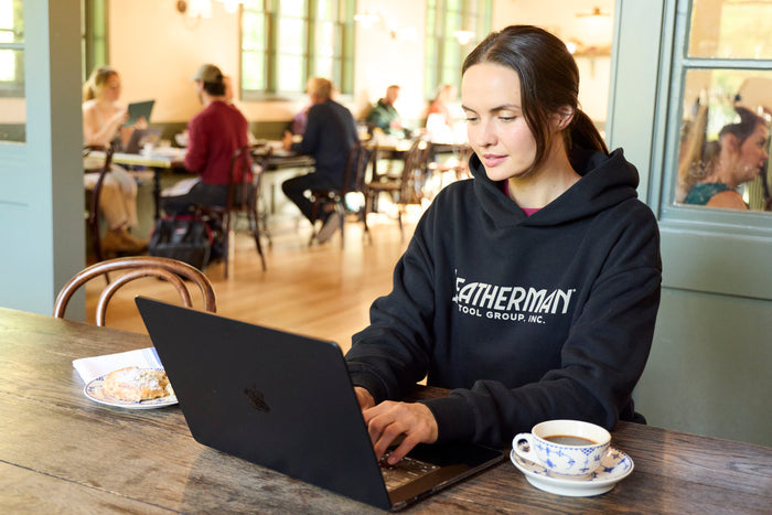 A woman sitting indoors working on a computer while wearing a black Leatherman Blade & Grain hoodie with a pastry and coffee on either side of the laptop.
