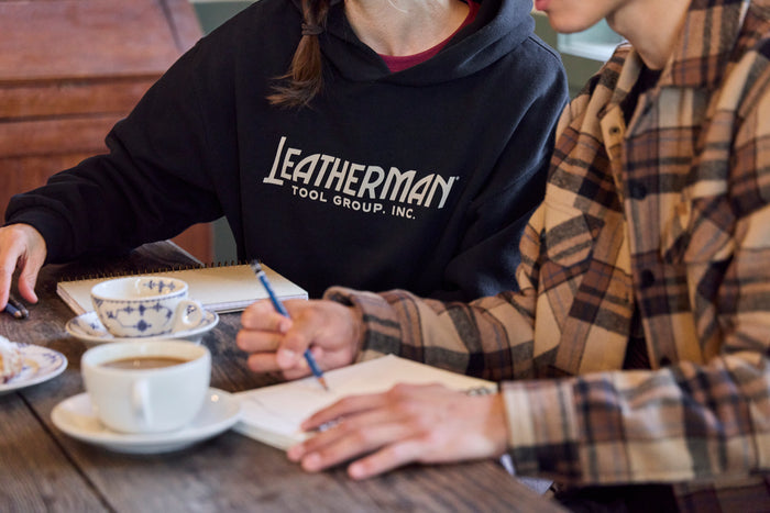 Two people sitting at a table with coffee, pastries and notebooks. One is wearing a black Leatherman Blade & Grain hoodie with 'Leatherman Tool Group Inc.' on the front.