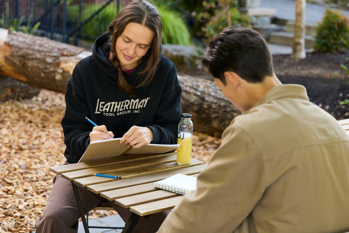 Two people sitting at a table outside with notebooks and pencils.. One is wearing a black Leatherman Blade & Grain Hoodie with 'Leatherman Tool Group Inc.' on the front.