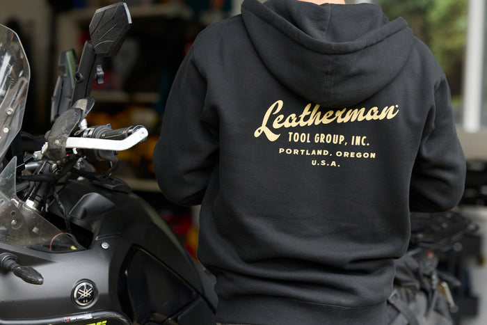 A person wearing a black Leatherman Vintage Zip-Up Hoodie standing next to a motorcycle, showing the gold Leatherman Tool Group, Inc. logo printed on the back.